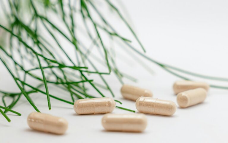 Een groep beige capsules ligt verspreid op een wit oppervlak, met lange, dunne, groene, grasachtige bladeren op de achtergrond.