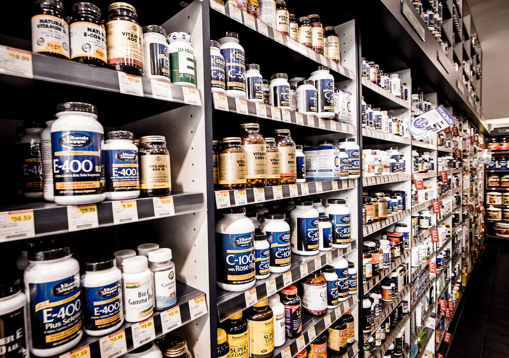 Planken vol met verschillende flessen vitaminen en supplementen, georganiseerd in rijen. De etiketten tonen verschillende merken en typen, zoals vitamine E en natuurlijke supplementen. Prijskaartjes zijn zichtbaar onder sommige producten.