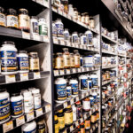 Planken vol met verschillende flessen vitaminen en supplementen, georganiseerd in rijen. De etiketten tonen verschillende merken en typen, zoals vitamine E en natuurlijke supplementen. Prijskaartjes zijn zichtbaar onder sommige producten.