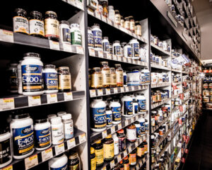 Planken vol met verschillende flessen vitaminen en supplementen, georganiseerd in rijen. De etiketten tonen verschillende merken en typen, zoals vitamine E en natuurlijke supplementen. Prijskaartjes zijn zichtbaar onder sommige producten.