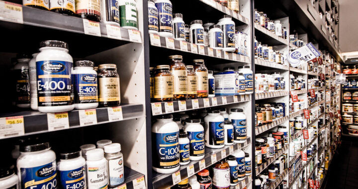 Planken vol met verschillende flessen vitaminen en supplementen, georganiseerd in rijen. De etiketten tonen verschillende merken en typen, zoals vitamine E en natuurlijke supplementen. Prijskaartjes zijn zichtbaar onder sommige producten.
