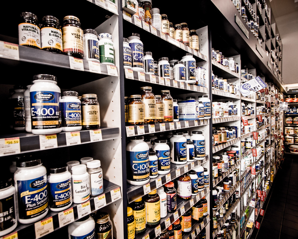 Planken vol met verschillende flessen vitaminen en supplementen, georganiseerd in rijen. De etiketten tonen verschillende merken en typen, zoals vitamine E en natuurlijke supplementen. Prijskaartjes zijn zichtbaar onder sommige producten.