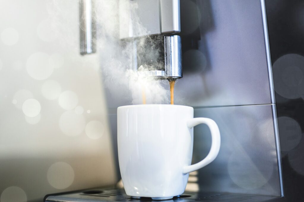 Een witte mok staat onder een espressomachine, die hete koffie schenkt. Stoom stijgt op rond de mok, wat de versheid en temperatuur van de koffie aangeeft. De achtergrond is zacht vervaagd, waardoor de aandacht op het brouwproces wordt gevestigd.