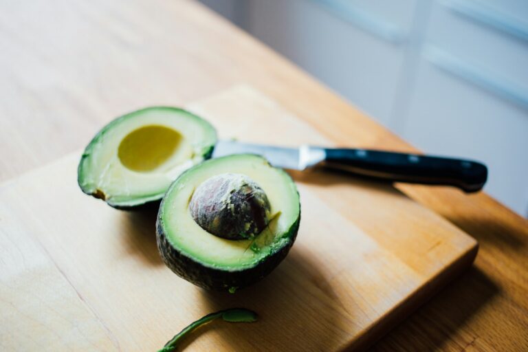 Een rijpe avocado in tweeën gesneden op een houten snijplank. Er ligt een mes met een zwart handvat naast en de pit is in één helft zichtbaar. De achtergrond toont een wazige keukensetting.