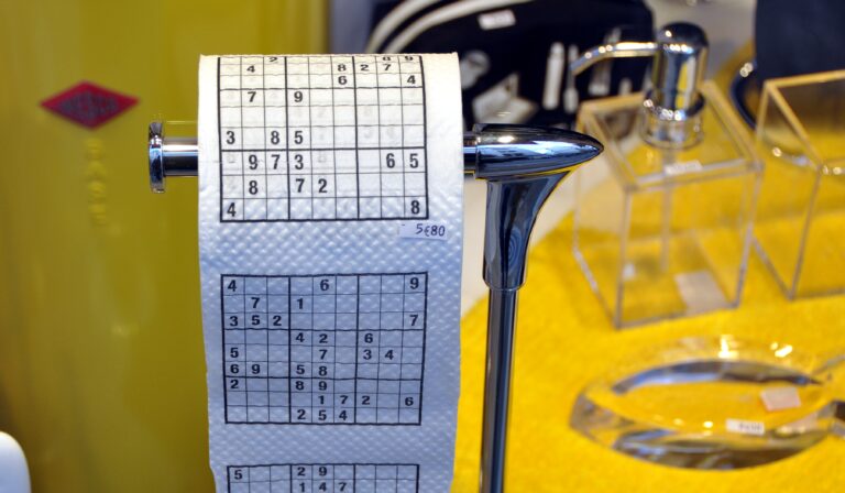 Een rol toiletpapier met Sudoku-puzzels erop gedrukt, wordt op een glimmende metalen houder geplaatst. De achtergrond omvat verschillende badkamerartikelen en accessoires in een winkeldisplay.
