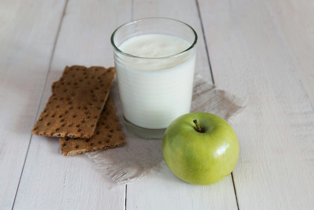 Een glas melk staat op een stoffen servet, naast twee rechthoekige crackers en een groene appel op een wit houten oppervlak.