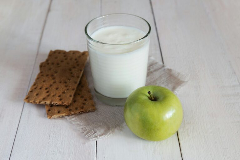 Een glas melk staat op een stoffen servet, naast twee rechthoekige crackers en een groene appel op een wit houten oppervlak.