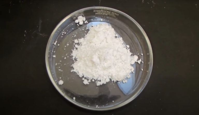 A clear glass petri dish contains a piled mound of white powder (magnesium carbonate). The dish is placed on a black surface, and small particles of the powder are scattered around it.