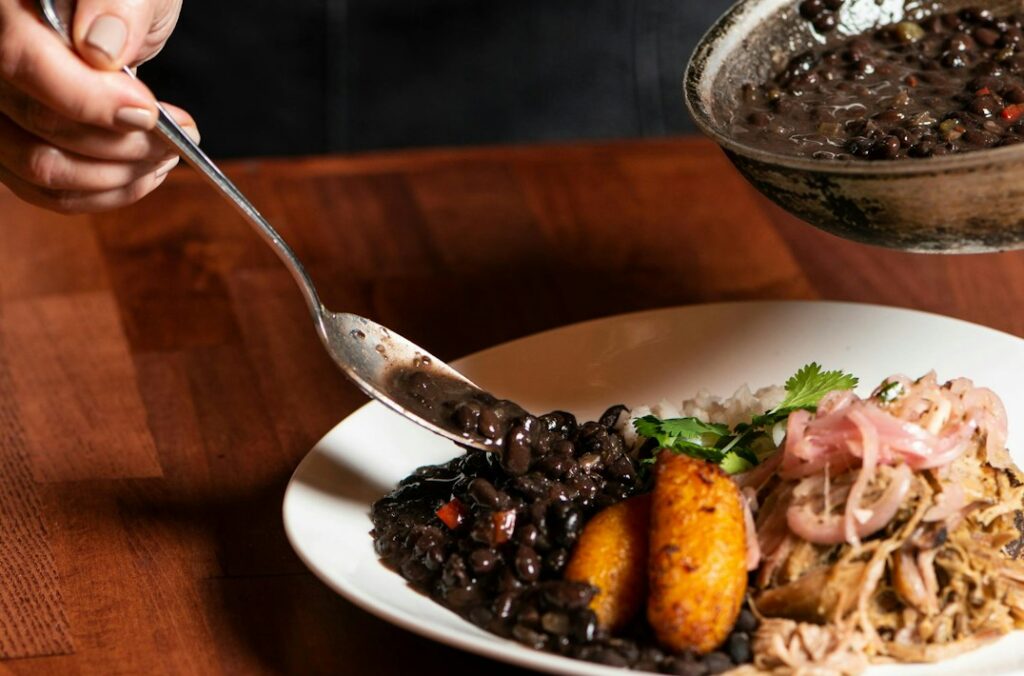 Een persoon serveert zwarte bonen uit een koekenpan op een bord. Het bord bevat rijst, pulled meat, gebakken bakbananen, ingelegde uien en koriander. De scène is op een houten tafel.