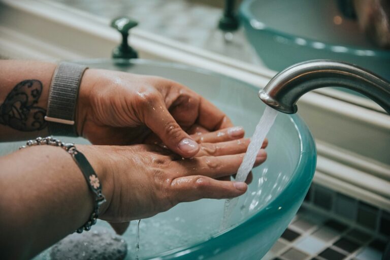 Twee handen worden gewassen onder stromend water in een glazen wasbak. De persoon heeft een armband en een tatoeage op zijn pols. De wasbak is geplaatst in een betegeld aanrecht en er stroomt water uit de kraan op de handen.