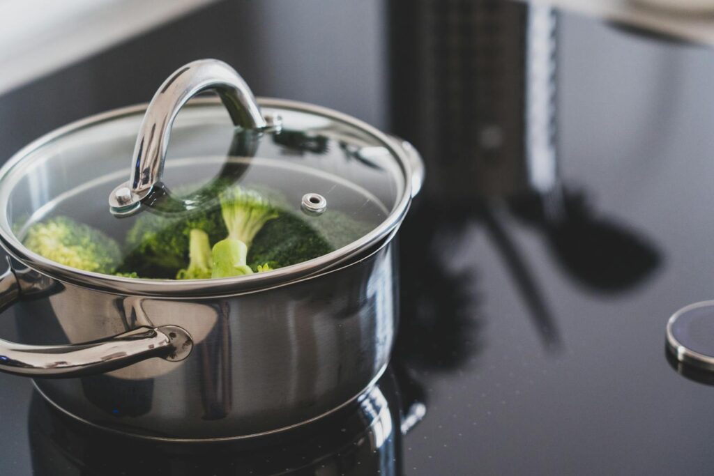 Een roestvrijstalen pot met een glazen deksel staat op een glazen fornuis. In de pot wordt broccoli gekookt. Op de achtergrond is een doosrasp gedeeltelijk zichtbaar.