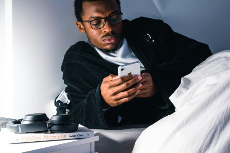 Een persoon met een bril ligt in bed en houdt een smartphone vast. Ze dragen een donkere jurk. Op het nachtkastje liggen een koptelefoon en een boek met de titel "Hábitos Atómicos" van James Clear.