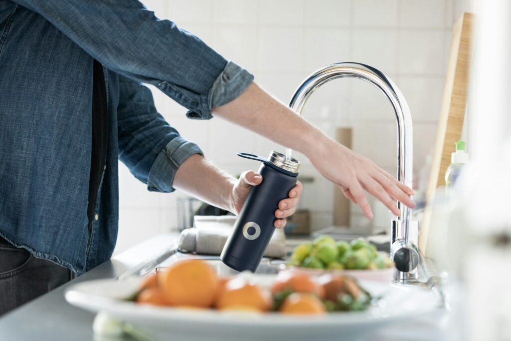 Een persoon in een spijkerblouse vult een donkerblauwe herbruikbare waterfles bij een gootsteen in de keuken. Op de voorgrond staat een fruitschaal met sinaasappels en citroenen. Op de achtergrond staan een betegelde muur en andere keukenspullen.