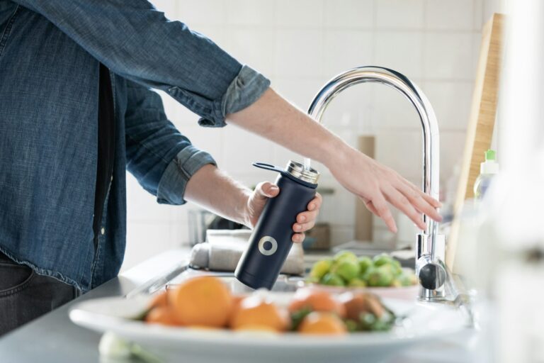 Een persoon in een spijkerblouse vult een donkerblauwe herbruikbare waterfles bij een gootsteen in de keuken. Op de voorgrond staat een fruitschaal met sinaasappels en citroenen. Op de achtergrond staan een betegelde muur en andere keukenspullen.