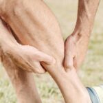 Een persoon die blauwe sportbroeken en hardloopschoenen draagt, is buiten en houdt zijn kuit met beide handen vast, alsof hij last heeft van ongemak of kramp. De scène suggereert dat hij misschien aan het stretchen is of een beenprobleem aanpakt.