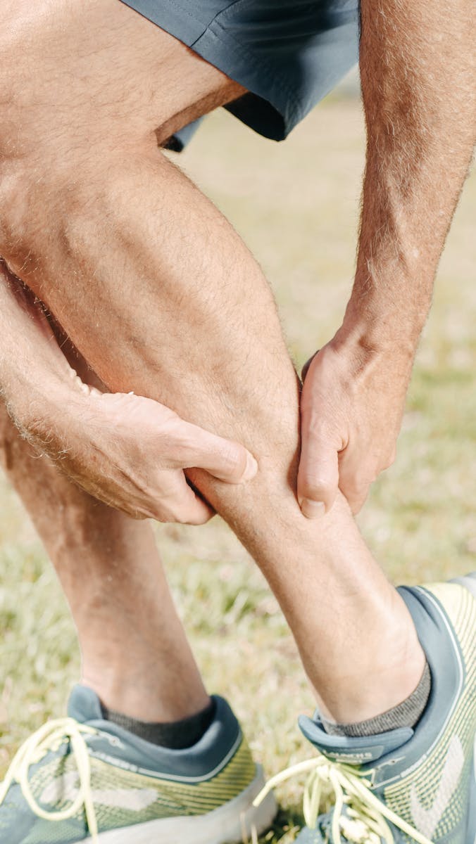 Een persoon die blauwe sportbroeken en hardloopschoenen draagt, is buiten en houdt zijn kuit met beide handen vast, alsof hij last heeft van ongemak of kramp. De scène suggereert dat hij misschien aan het stretchen is of een beenprobleem aanpakt.