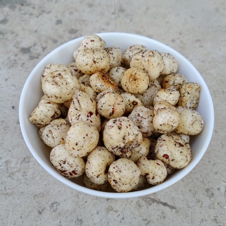 Een witte kom gevuld met gepofte lotuszaden (Makhana) staat op een getextureerd betonnen oppervlak. De zaden zijn wit met een gevlekt uiterlijk, met kleine bruine vlekjes.