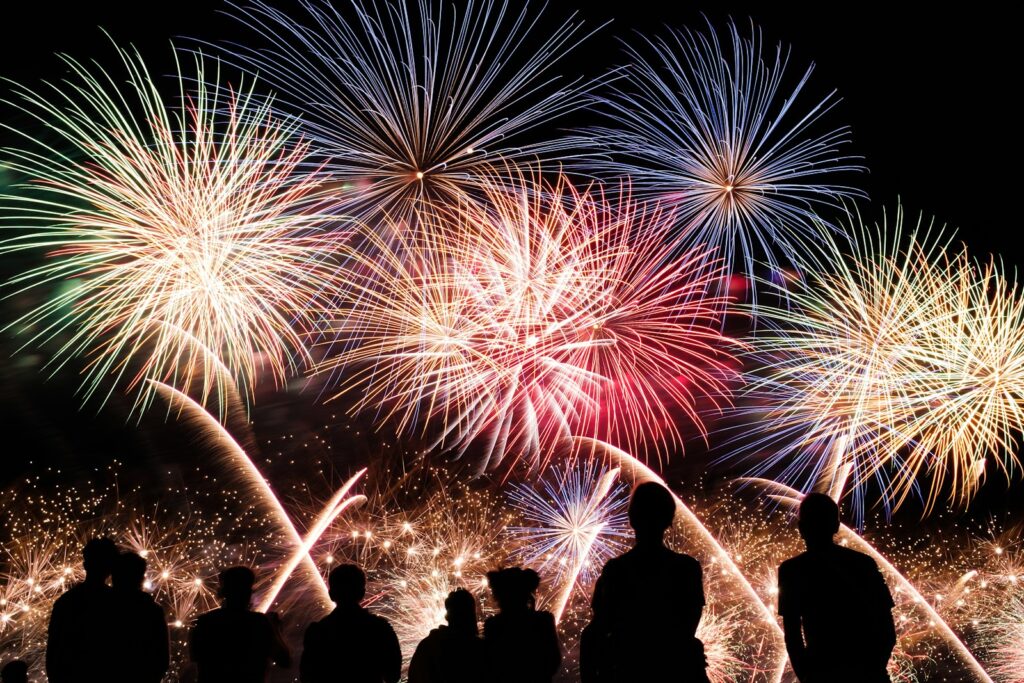 Silhouetten van mensen die kijken naar een levendig vuurwerkspektakel in de nachtelijke hemel. Meerdere uitbarstingen van kleurrijk vuurwerk, in tinten blauw, rood en geel, verlichten de scène en creëren een feestelijke sfeer.