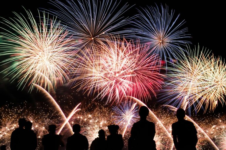 Silhouetten van mensen die kijken naar een levendig vuurwerkspektakel in de nachtelijke hemel. Meerdere uitbarstingen van kleurrijk vuurwerk, in tinten blauw, rood en geel, verlichten de scène en creëren een feestelijke sfeer.