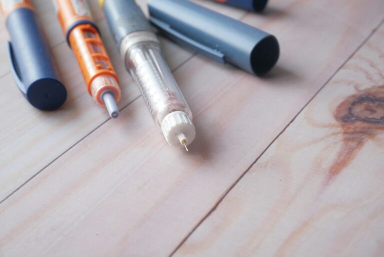 Drie insulinepennen liggen op een houten oppervlak met hun doppen verwijderd, waardoor de naalden zichtbaar zijn. De pennen hebben verschillende kleuren: oranje, blauw en transparant. Doppen zijn zichtbaar naast de pennen.