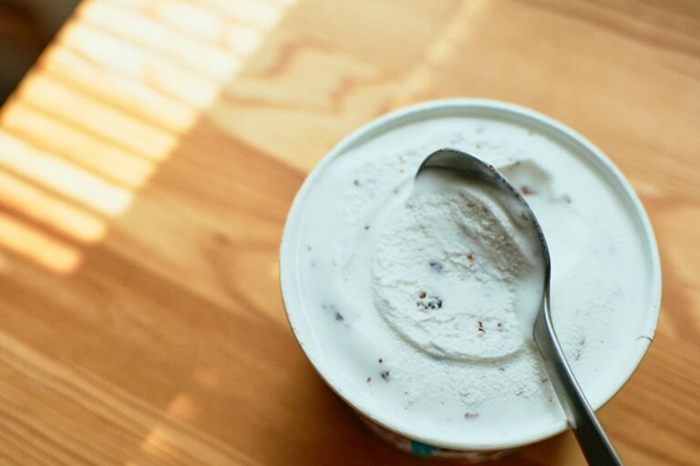 Een bak vanille-ijs met chocoladechips staat gedeeltelijk open op een houten tafel. Een metalen lepel rust op het ijs. Zonlicht werpt schaduwen op het tafeloppervlak.