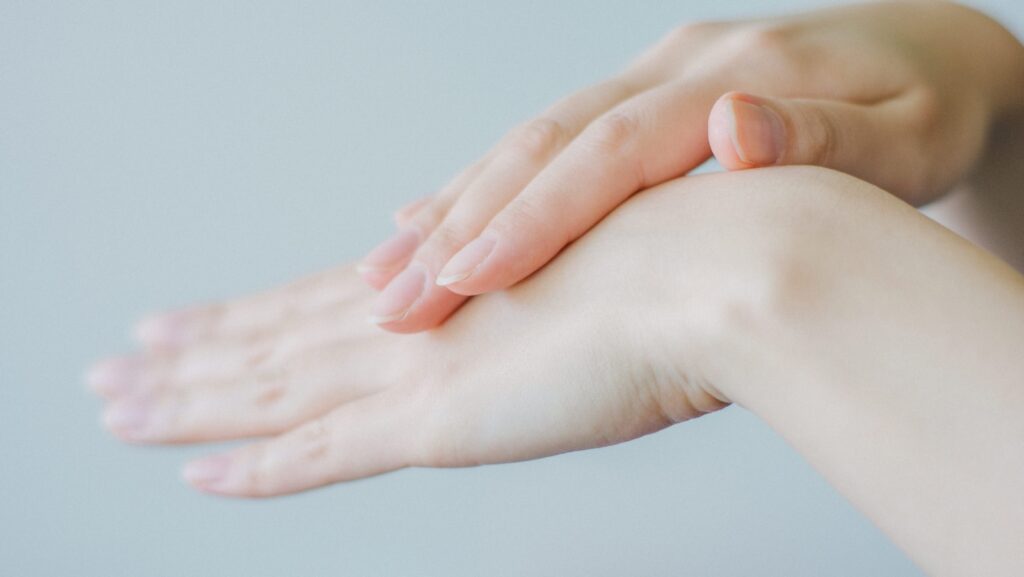 Close-up of a pair of hands. One hand is placed gently on top of the other against a blurred light background. The nails are neatly manicured.