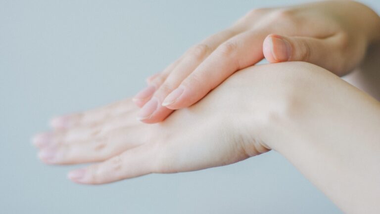 Close-up of a pair of hands. One hand is placed gently on top of the other against a blurred light background. The nails are neatly manicured.