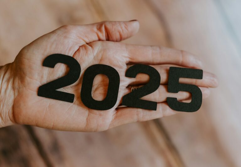 Een hand houdt zwarte uitgesneden cijfers vast die zo zijn gerangschikt dat ze "2025" vormen, tegen een wazige houten achtergrond.