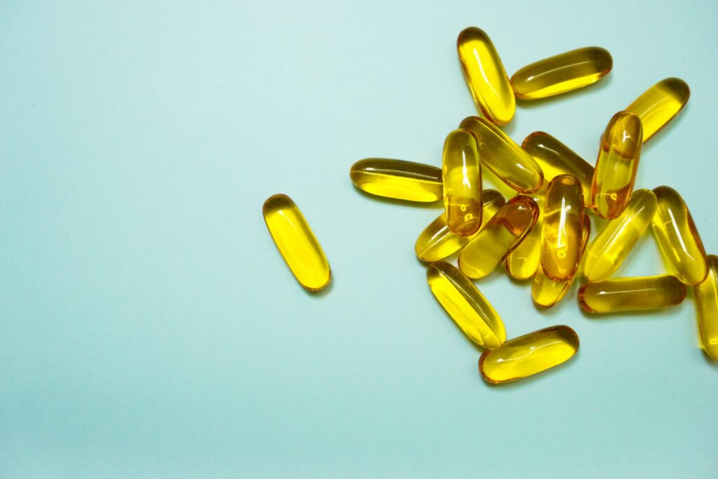 Een groep gele, doorschijnende gelcapsules ligt verspreid op een lichtblauw oppervlak, waarbij één capsule gescheiden is van de hoofdcluster. Viamine D