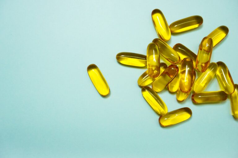 Een groep gele, doorschijnende gelcapsules ligt verspreid op een lichtblauw oppervlak, waarbij één capsule gescheiden is van de hoofdcluster. Viamine D