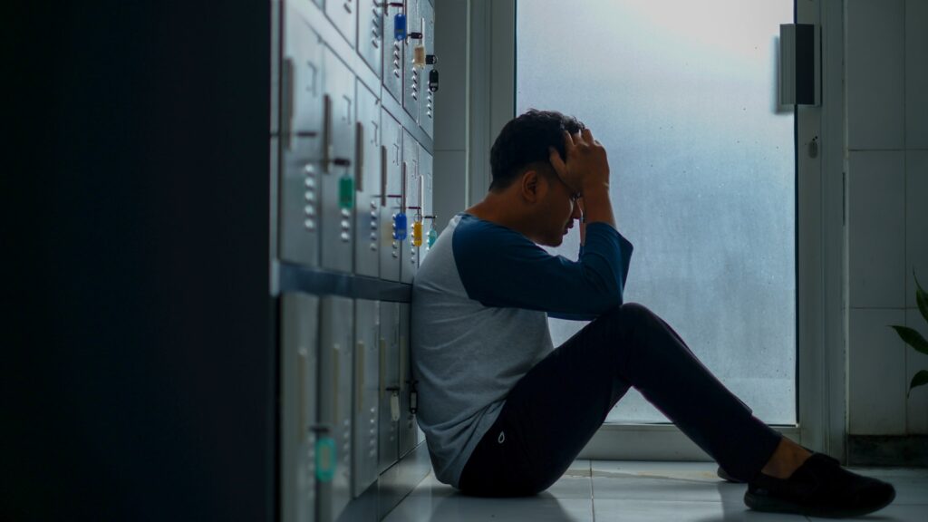 Een persoon zit op de grond tegen een kluisje en houdt zijn hoofd met beide handen vast. De scène speelt zich binnen af, vlakbij een deur met matglas, en de houding van de persoon suggereert stress of stress.