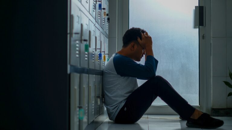 Een persoon zit op de grond tegen een kluisje en houdt zijn hoofd met beide handen vast. De scène speelt zich binnen af, vlakbij een deur met matglas, en de houding van de persoon suggereert stress of stress.