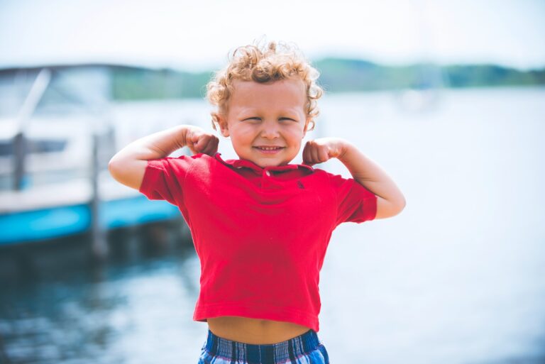 Een jong kind met krullend haar, gekleed in een rood shirt en een geruite korte broek, staat buiten bij het water en lacht met beide armen naar de camera op een zonnige dag.