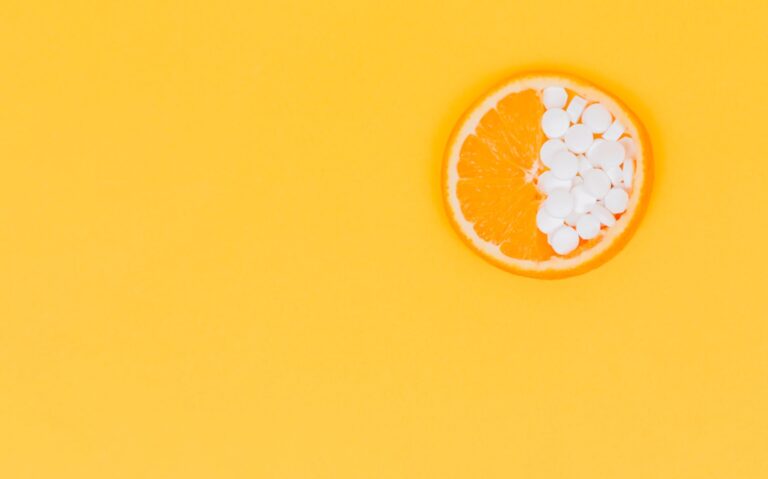Een schijfje sinaasappel met daarop een aantal kleine, witte, ronde tabletten, tegen een effen gele achtergrond.