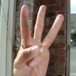 Voor een bakstenen muur is een hand te zien die drie vingers omhoog houdt. Op de achtergrond staan een raam met uitzicht op de straat en wat tuinmeubilair.