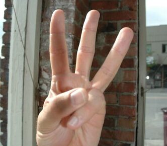 Voor een bakstenen muur is een hand te zien die drie vingers omhoog houdt. Op de achtergrond staan een raam met uitzicht op de straat en wat tuinmeubilair.