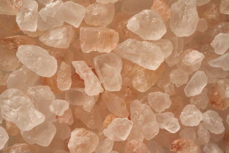 Close-up van verschillende onregelmatig gevormde roze Himalaya-zoutkristallen, variërend in kleur van wit tot lichtroze en oranje, die samenkomen tegen een neutrale achtergrond.