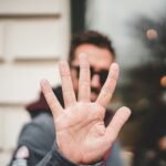 Een persoon houdt een open hand dicht bij de camera, waardoor zijn gezicht niet zichtbaar is. De achtergrond is onscherp en toont een deel van een gebouw en wat reflecties in een raam.