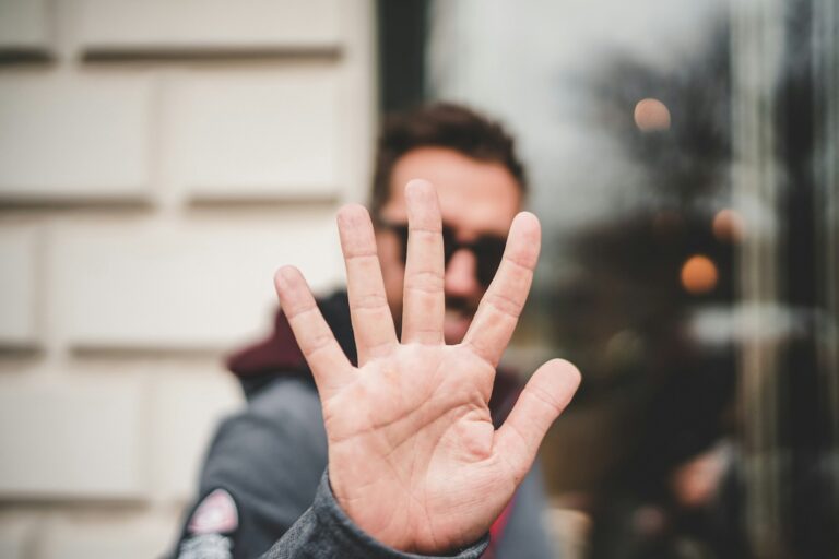 Een persoon houdt een open hand dicht bij de camera, waardoor zijn gezicht niet zichtbaar is. De achtergrond is onscherp en toont een deel van een gebouw en wat reflecties in een raam.