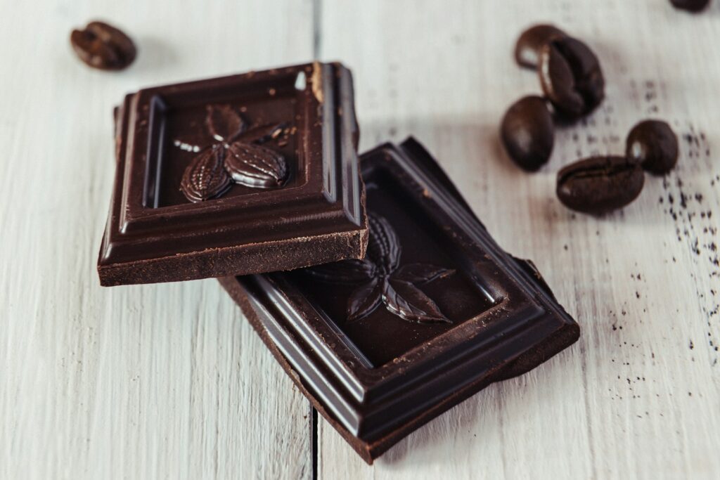 Twee stukjes pure chocolade met een cacaovruchtmotief liggen op een wit houten oppervlak, met enkele koffiebonen eromheen verspreid.