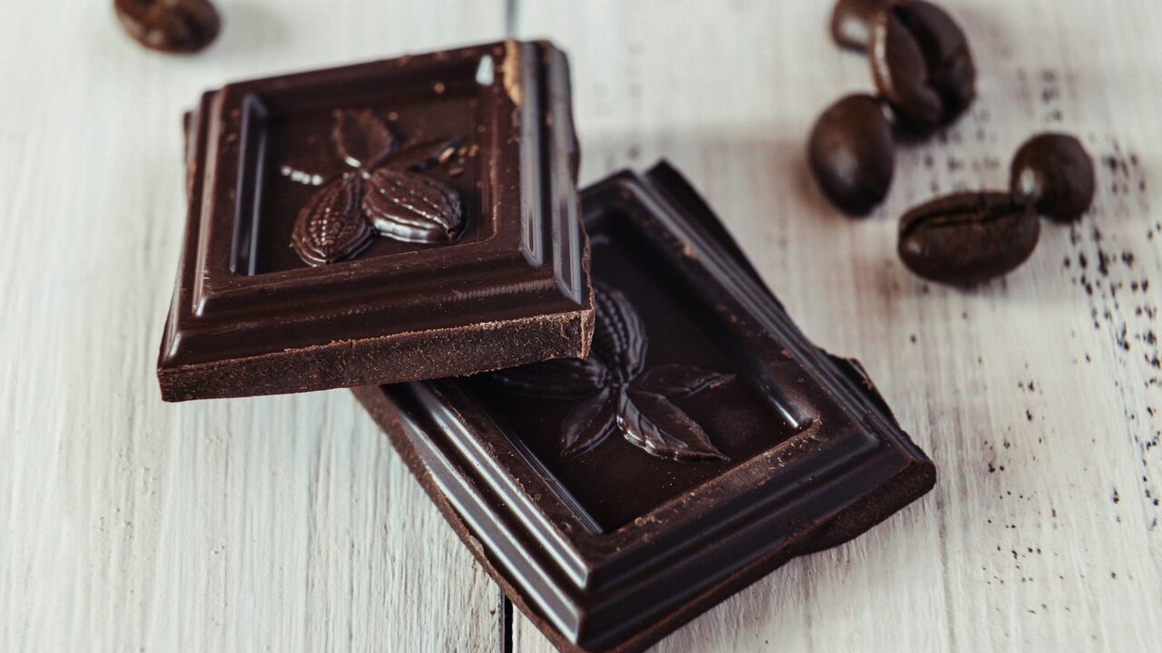 Twee stukjes pure chocolade met een cacaovruchtmotief liggen op een wit houten oppervlak, met enkele koffiebonen eromheen verspreid.