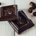 Twee stukjes pure chocolade met een cacaovruchtmotief liggen op een wit houten oppervlak, met enkele koffiebonen eromheen verspreid.