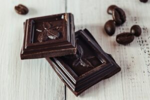 Twee stukjes pure chocolade met een cacaovruchtmotief liggen op een wit houten oppervlak, met enkele koffiebonen eromheen verspreid.