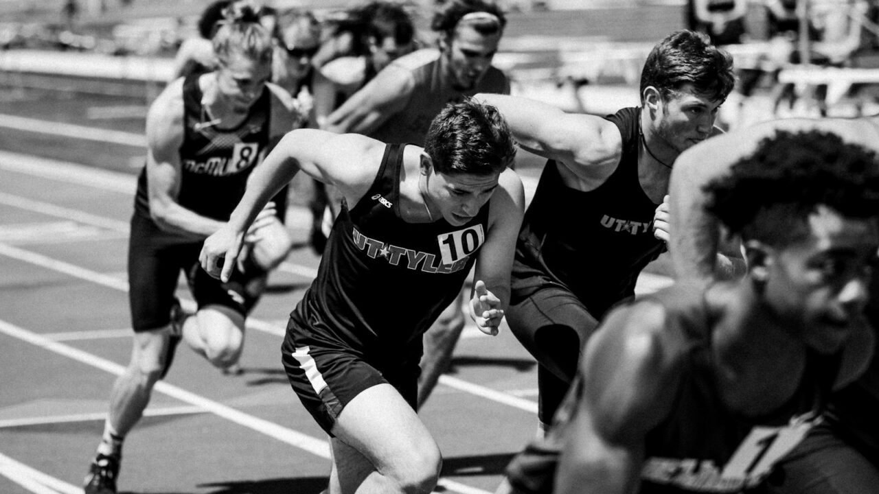 Een groep mannelijke atleten in hardloopkleding doet mee aan een sprintrace op een atletiekbaan, vastgelegd tijdens hun pas. De afbeelding is in zwart-wit.