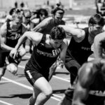 Een groep mannelijke atleten in hardloopkleding doet mee aan een sprintrace op een atletiekbaan, vastgelegd tijdens hun pas. De afbeelding is in zwart-wit.
