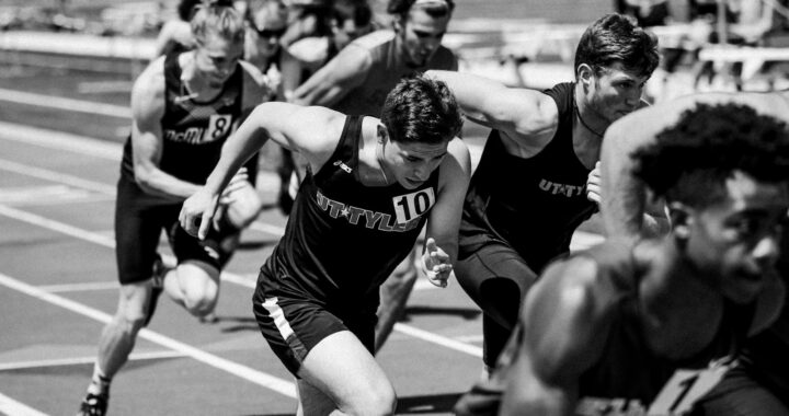 Een groep mannelijke atleten in hardloopkleding doet mee aan een sprintrace op een atletiekbaan, vastgelegd tijdens hun pas. De afbeelding is in zwart-wit.