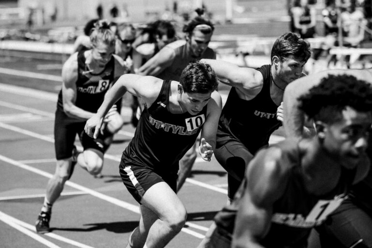 Een groep mannelijke atleten in hardloopkleding doet mee aan een sprintrace op een atletiekbaan, vastgelegd tijdens hun pas. De afbeelding is in zwart-wit.