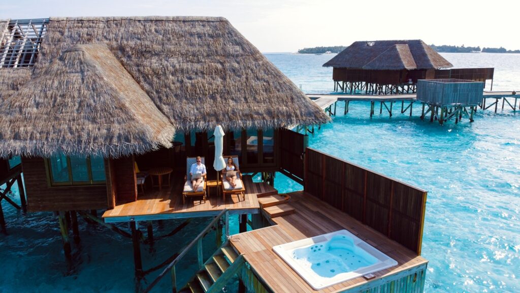 Een houten villa boven het water met een rieten dak torent hoog uit boven het helderblauwe water. Twee mensen ontspannen op ligstoelen op het terras bij een witte parasol, met een jacuzzi in de buurt en een andere villa zichtbaar op de achtergrond. Maladiven