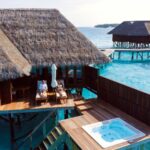 Een houten villa boven het water met een rieten dak torent hoog uit boven het helderblauwe water. Twee mensen ontspannen op ligstoelen op het terras bij een witte parasol, met een jacuzzi in de buurt en een andere villa zichtbaar op de achtergrond. Maladiven
