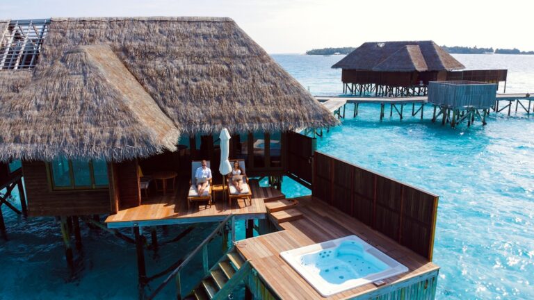 Een houten villa boven het water met een rieten dak torent hoog uit boven het helderblauwe water. Twee mensen ontspannen op ligstoelen op het terras bij een witte parasol, met een jacuzzi in de buurt en een andere villa zichtbaar op de achtergrond. Maladiven